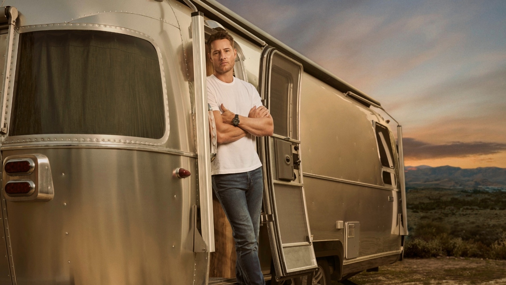 Man in a white t-shirt leans against a silver Airstream trailer at sunset, with a scenic background of hills and sky.