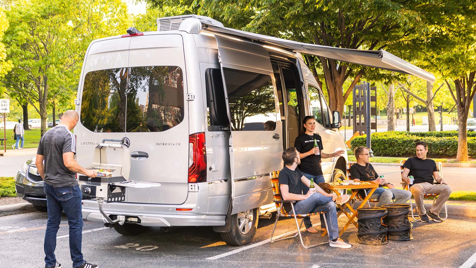 A group of people enjoying a barbecue and drinks by a parked camper van with an awning, surrounded by trees.