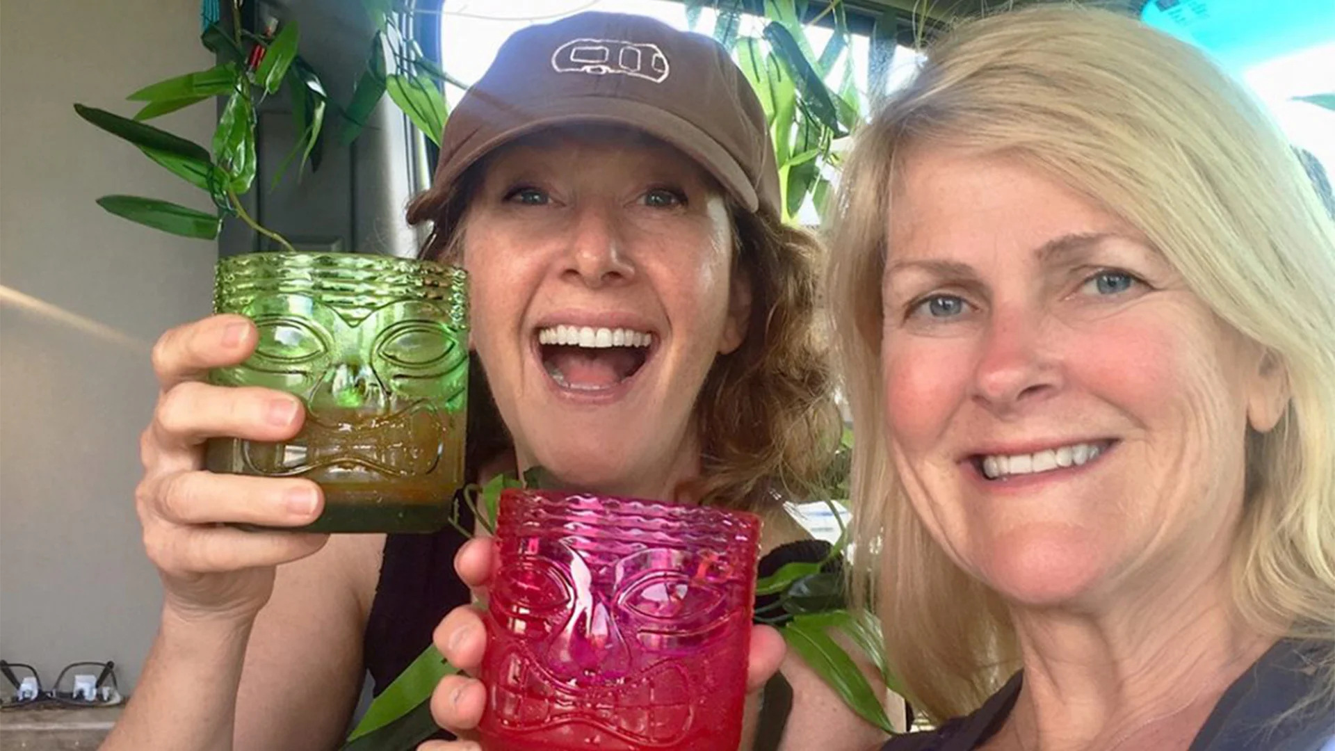 Two smiling women holding colorful tiki glasses, surrounded by green plants.