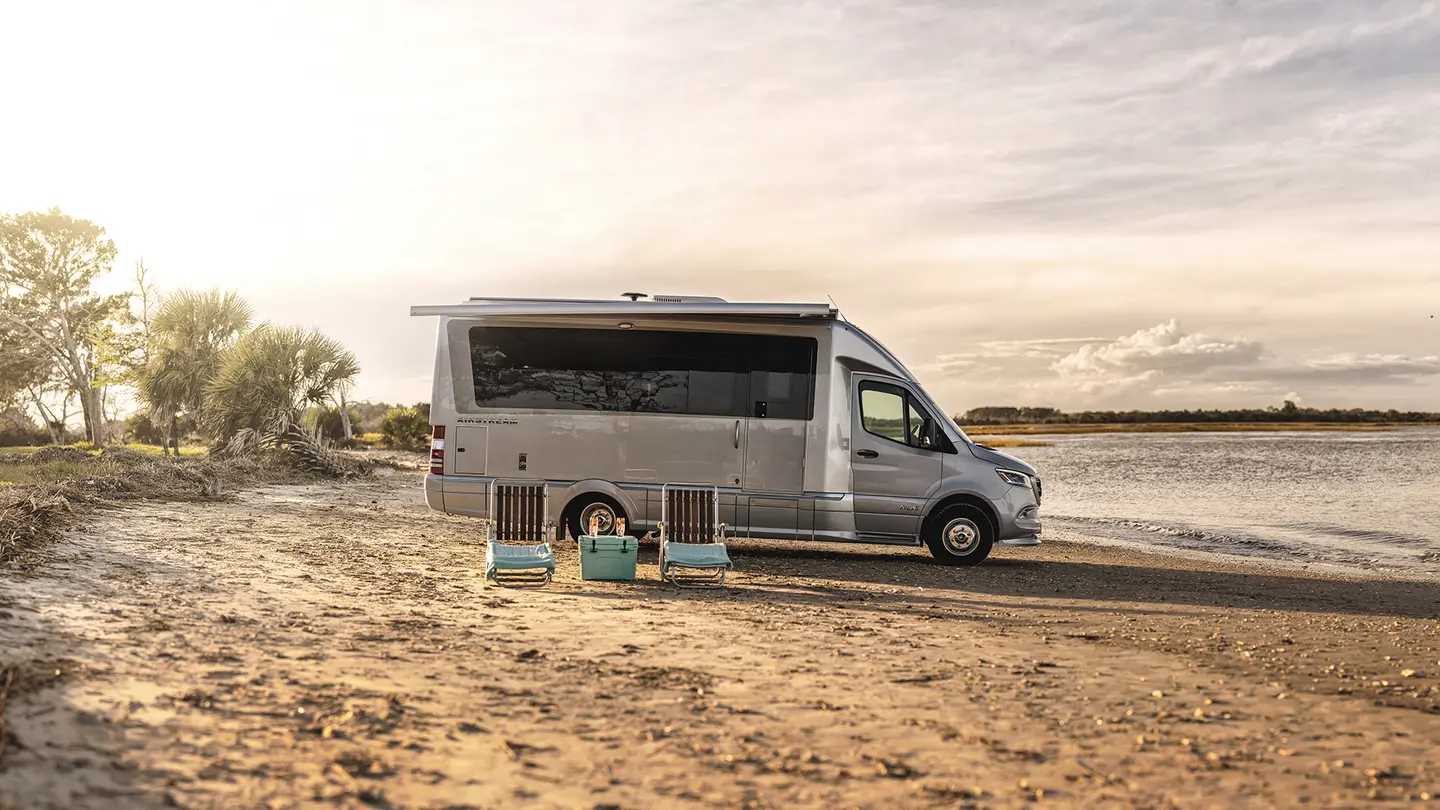 A silver camper van is parked on a sandy beach near the water. Four striped chairs and a cooler are arranged in front of the van, facing the shoreline. Trees and a cloudy sky are visible in the background.