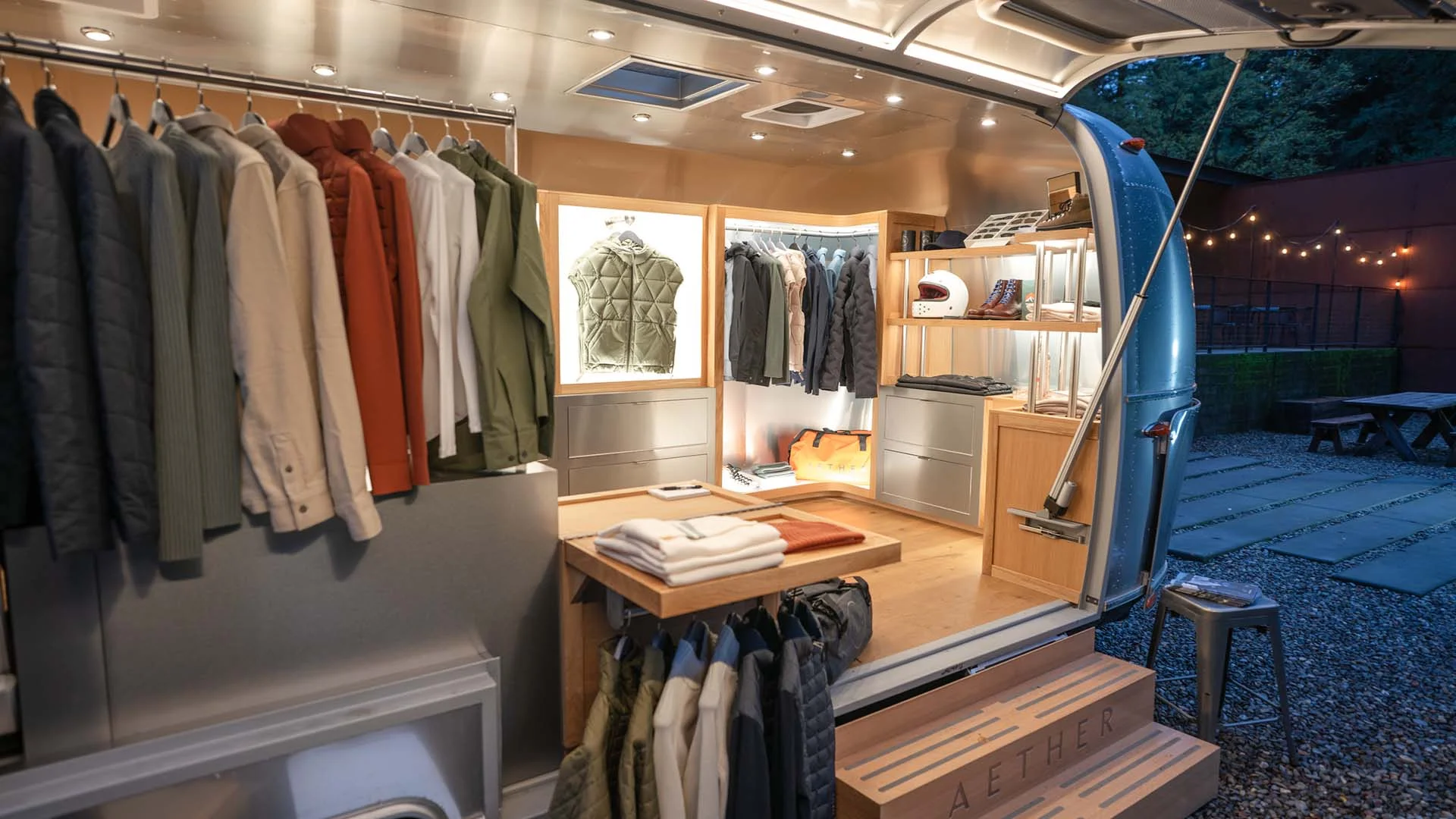 Interior of a mobile clothing boutique with jackets, helmets, and boots displayed inside a well-lit trailer on a gravel lot.