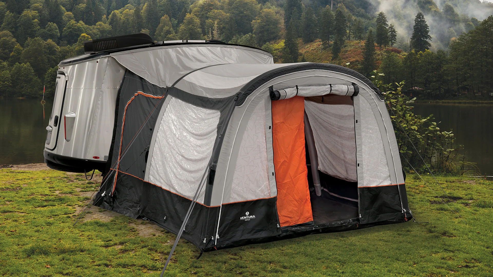 A gray and orange tent is attached to a small silver camper trailer on grassy ground near a lake, with forested hills and mist in the background.