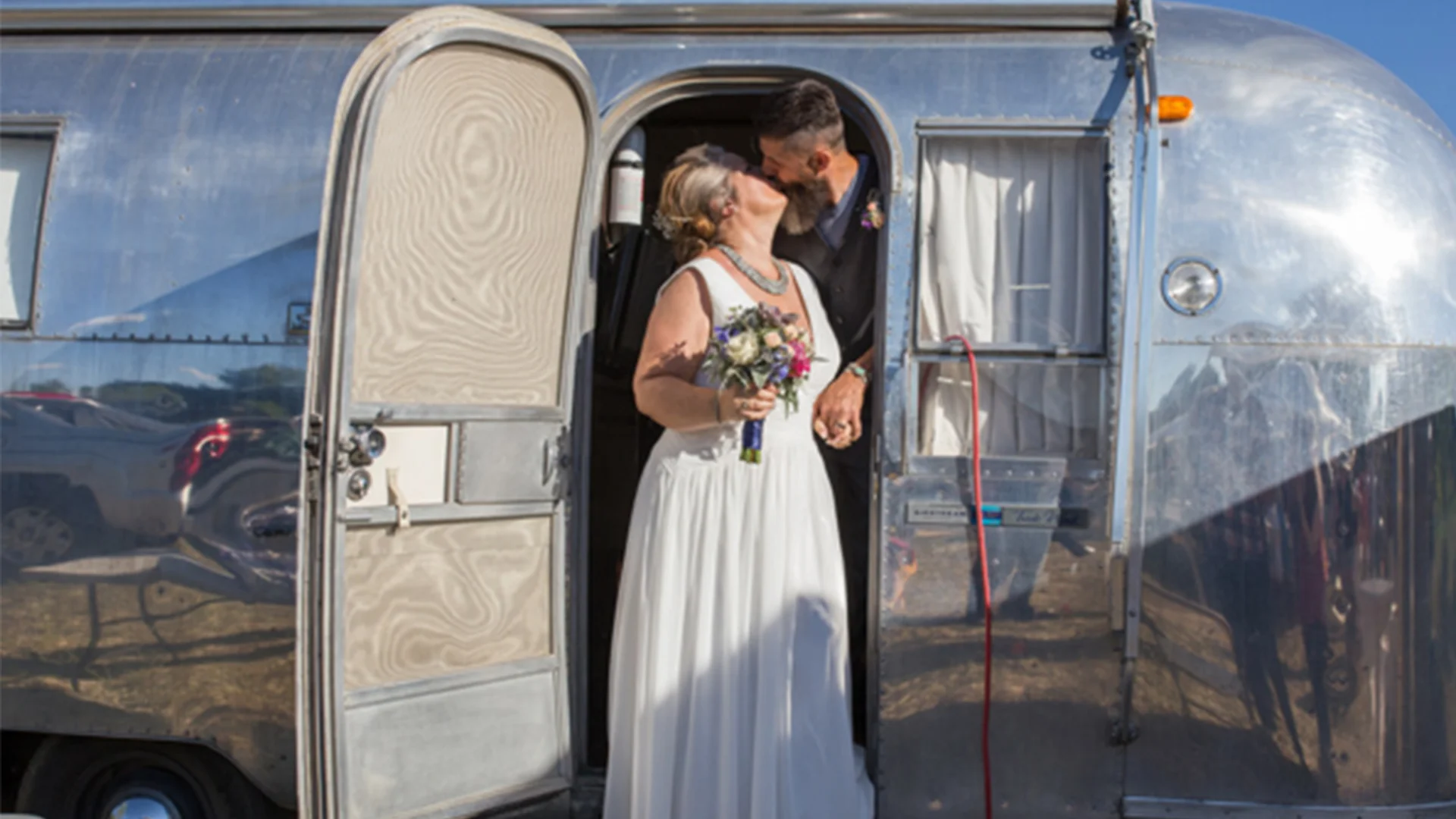 Bride and groom kiss at the doorway of a vintage silver trailer, with the bride holding a bouquet and wearing a white dress.