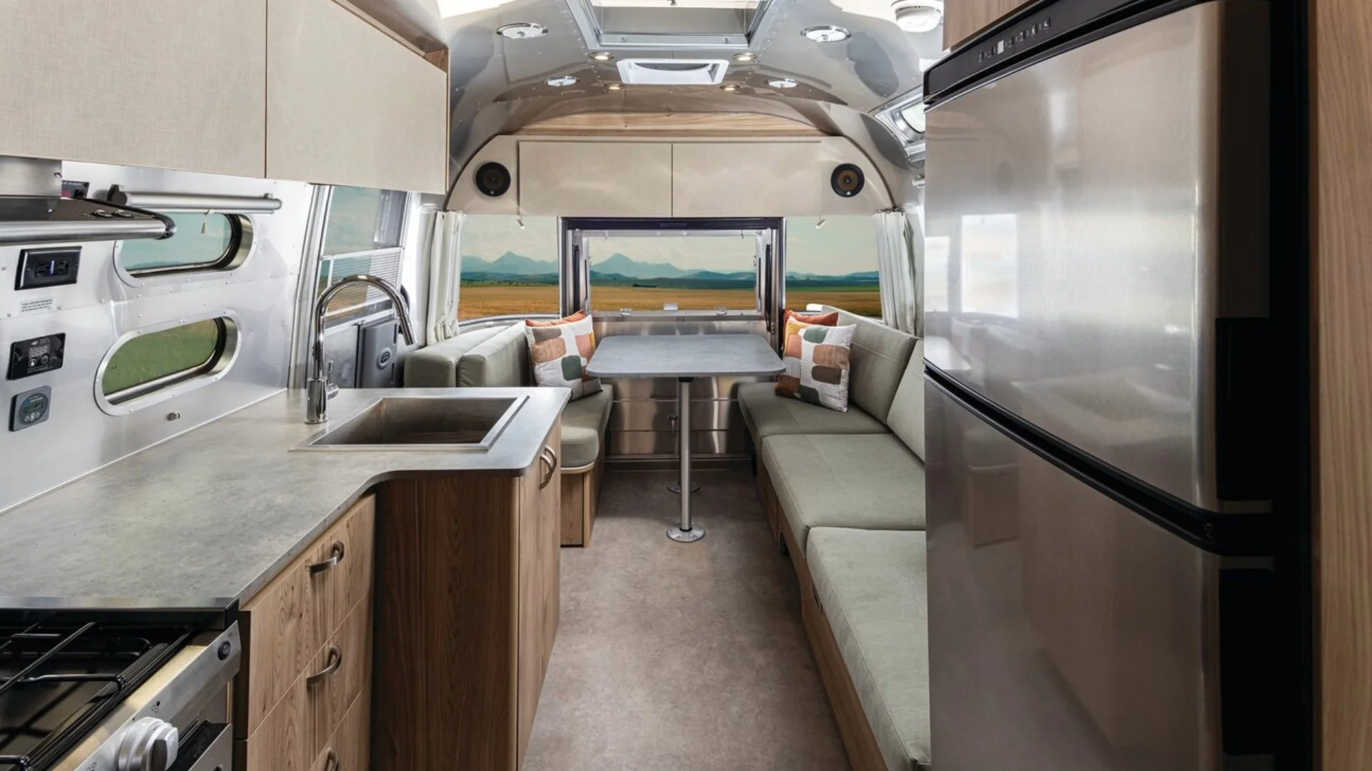 Interior of a modern travel trailer with a kitchen, small dining area, and large window view of mountains and fields.
