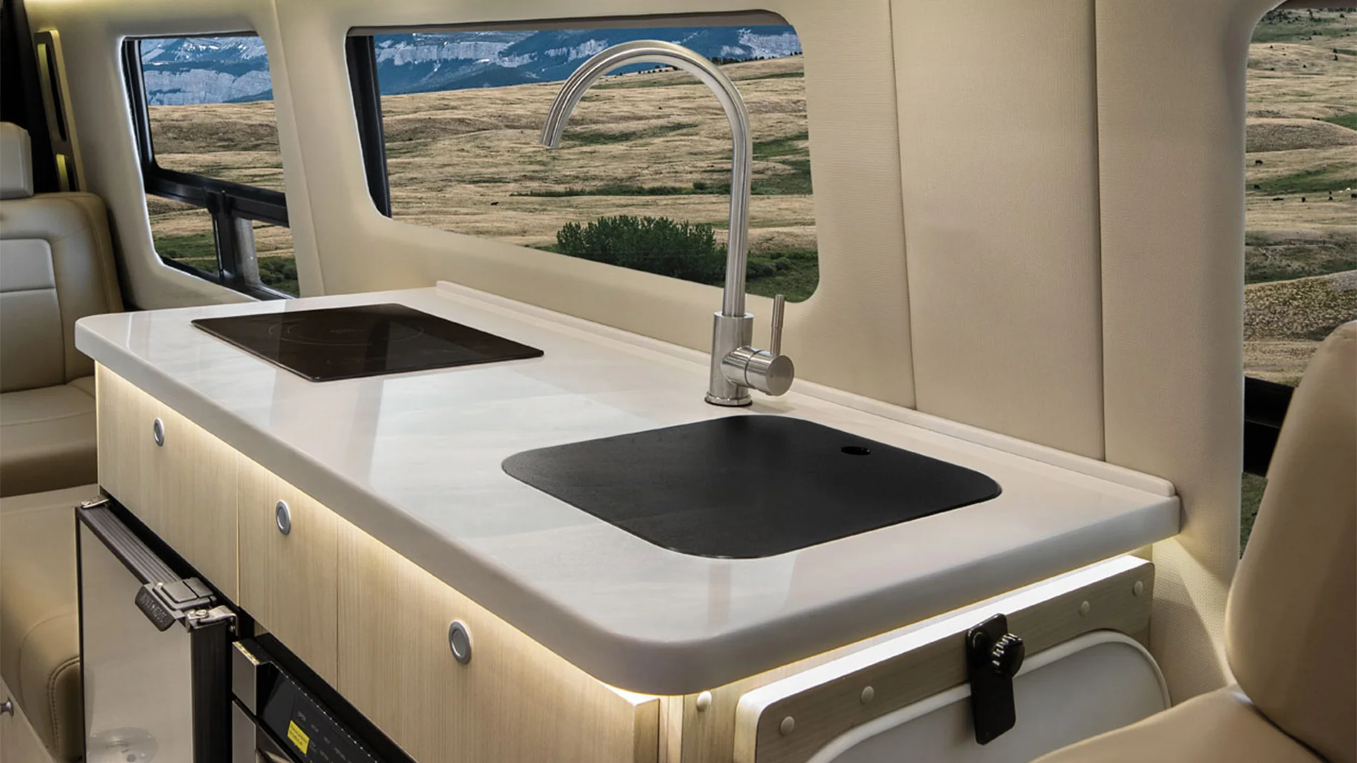 Modern van kitchen with a sleek countertop, sink, and stove. Large windows showcase a scenic landscape of rolling hills.