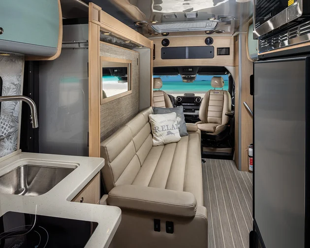 Interior of a modern RV with a kitchenette, sink, induction cooktop, beige leather couch, and driver’s seat, featuring a beach view outside.