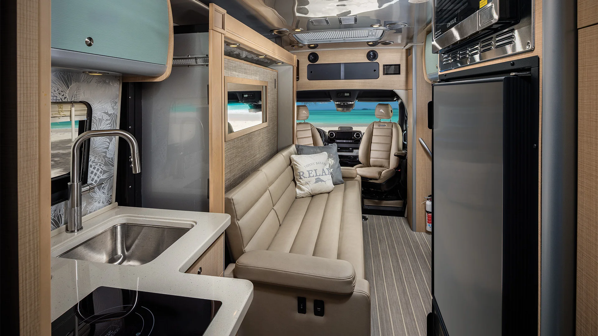 Interior of a modern RV with a kitchenette, sink, induction cooktop, beige leather couch, and driver’s seat, featuring a beach view outside.