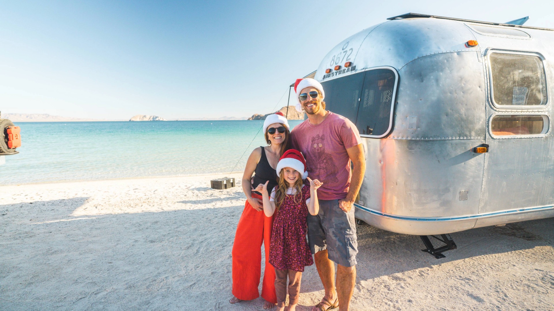 Family in Santa hats poses by an Airstream trailer on a sunny beach, with clear blue ocean in the background.