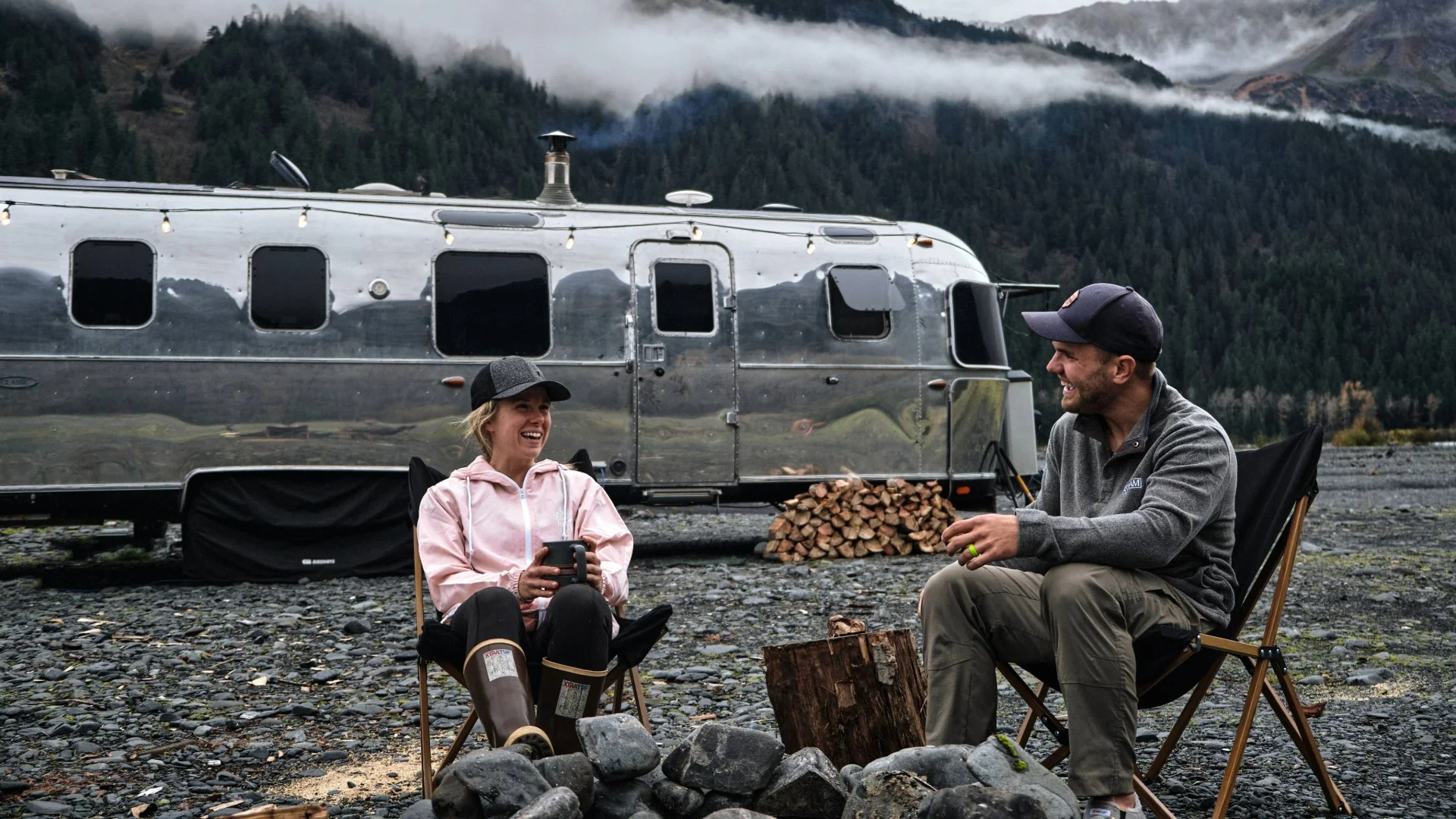 Two people sit laughing by a fire pit in front of an Airstream trailer, with a mountain backdrop and fog.