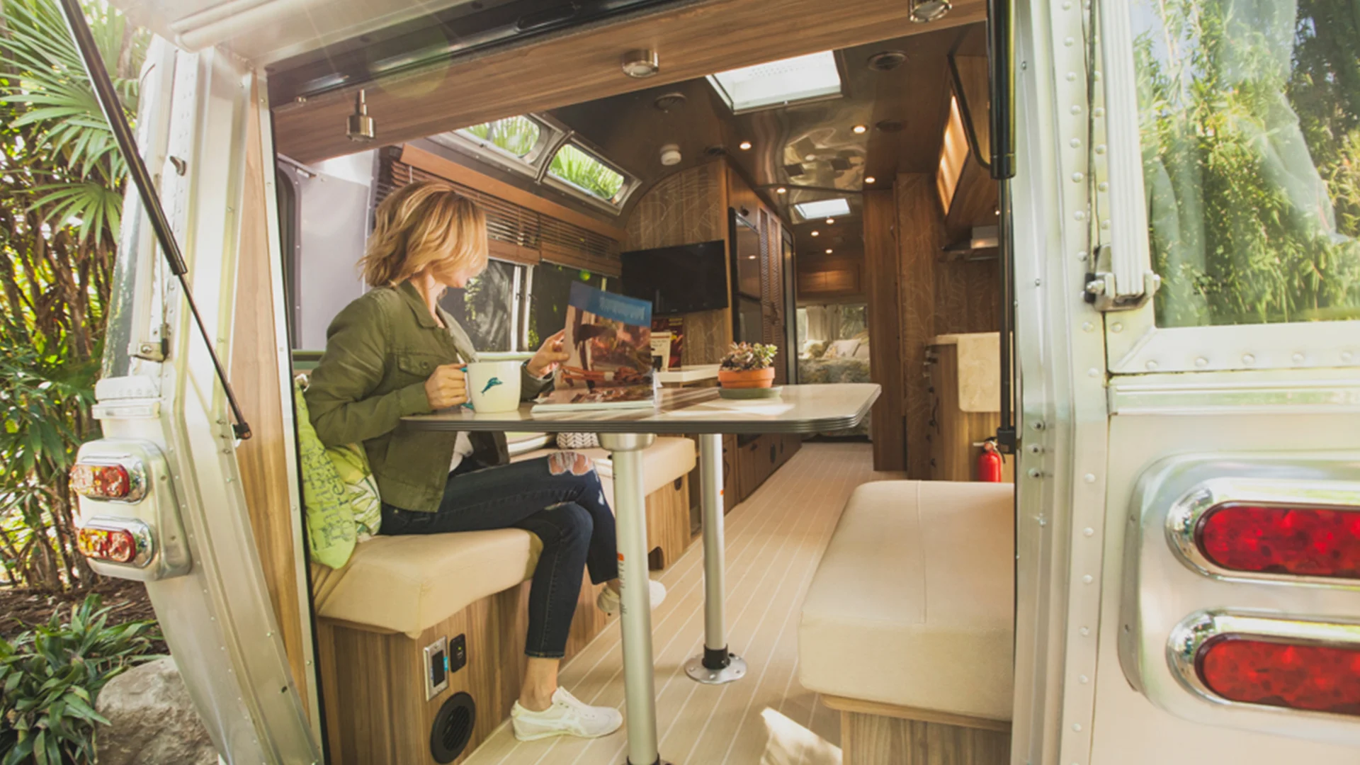 A woman sits inside a modern camper van, holding a mug and reading a magazine at a fold-out table. The van has wood-paneled interiors, natural light, and a lush outdoor setting visible through the open door.