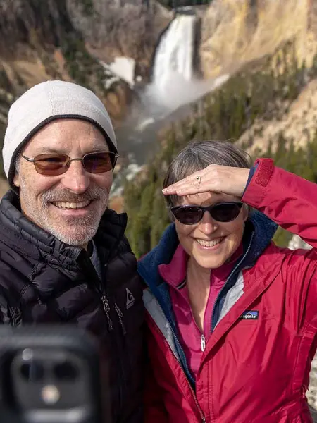 A man and woman smiling for a selfie with a waterfall and forested canyon in the background.