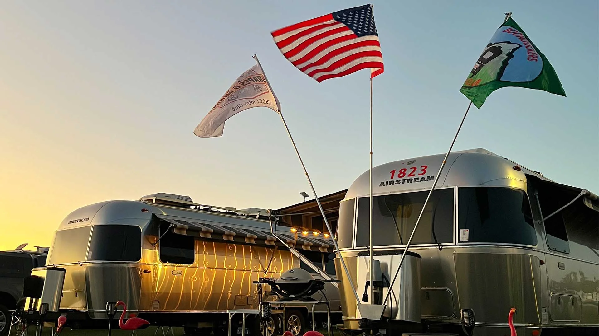 Silver Airstream trailers with flags, including an American flag, flutter in the wind at sunset, surrounded by pink flamingo decorations.