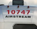Close-up of a silver Airstream trailer with "10747 Airstream" in red and black letters. A vent and sky are visible above.