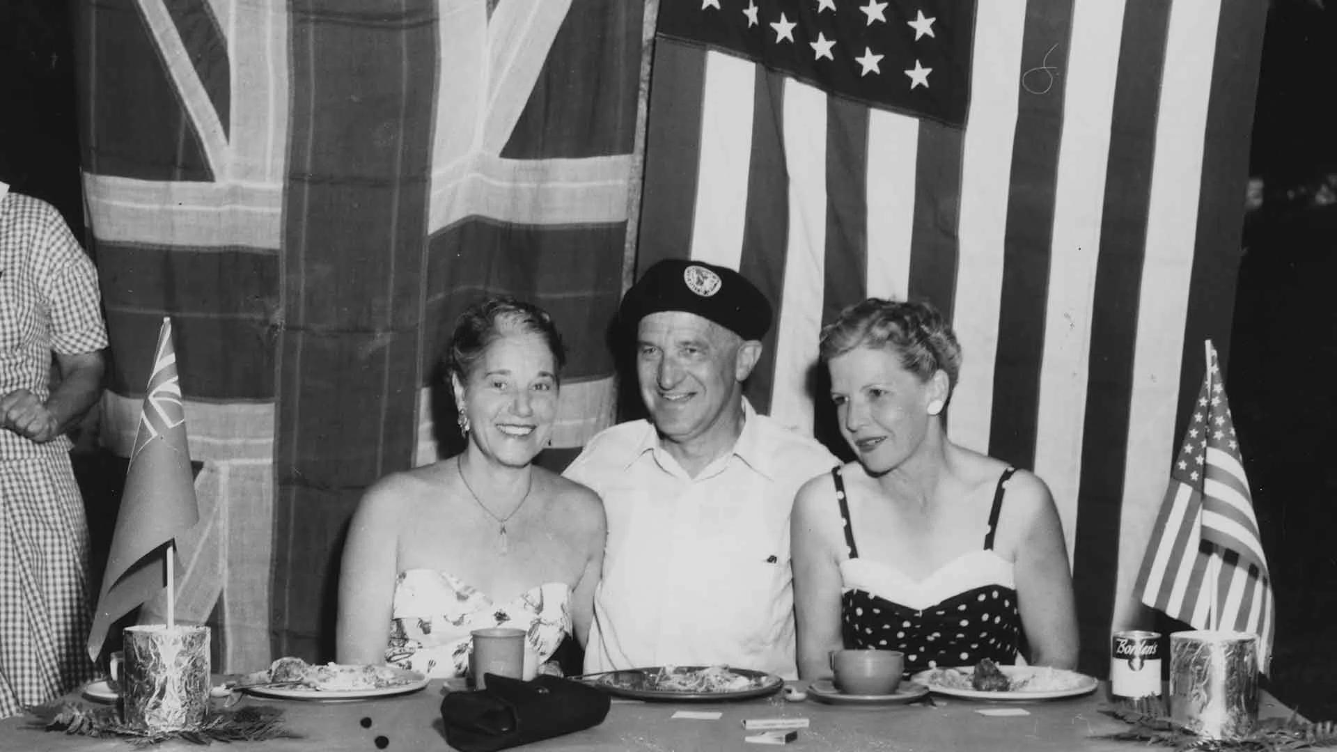 Three people sit at a table with food, in front of UK and US flags. A man in a beret is between two women. Small flags and plates are on the table.
