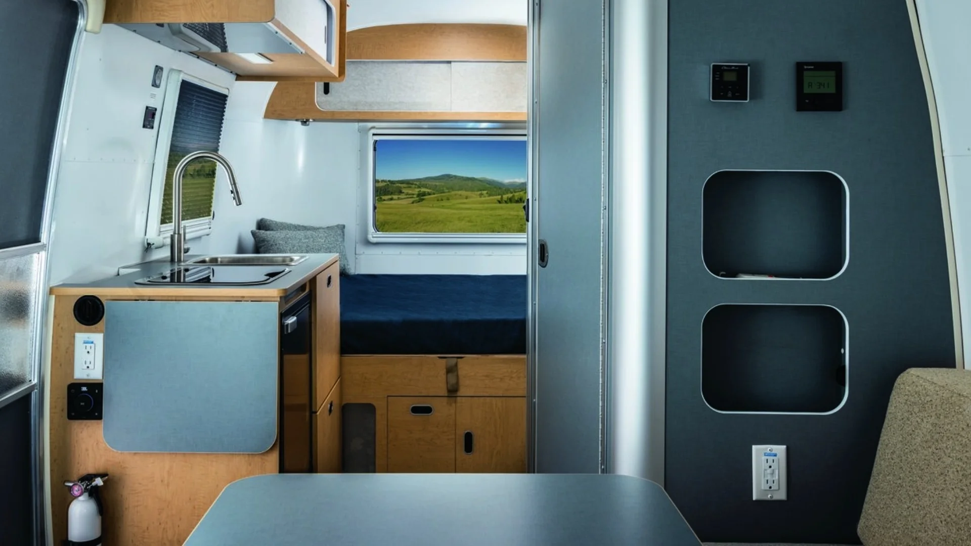 Interior of a modern camper van featuring a kitchen area with a sink, stovetop, bed, and a window with a view of green hills and blue sky.