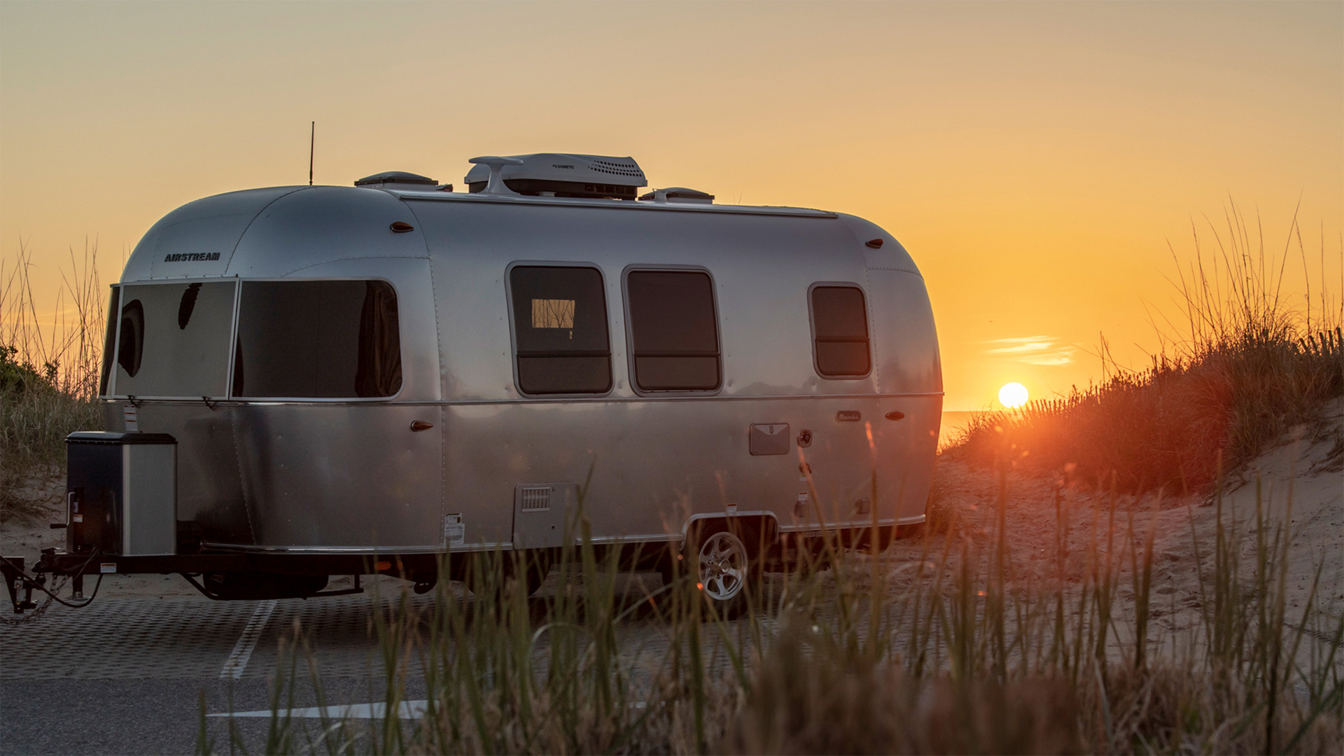 Silver camper trailer parked near dunes at sunset, with a warm glow illuminating the sky and grass in the foreground.