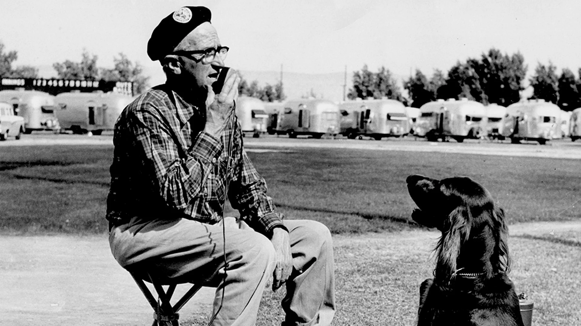 An older man in a plaid shirt and beret sits on a stool smoking a pipe, facing a dog, with a row of trailers in the background.