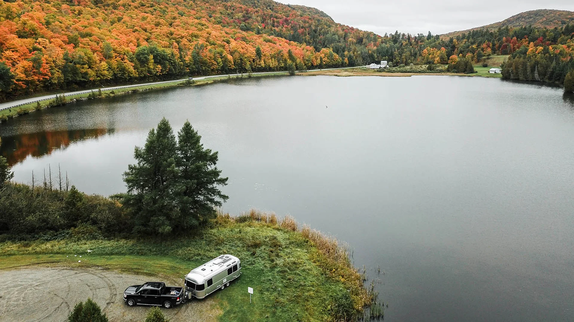 A truck towing a silver camper is parked by a calm lake surrounded by autumn trees with vibrant orange and green foliage, under an overcast sky in a hilly landscape.