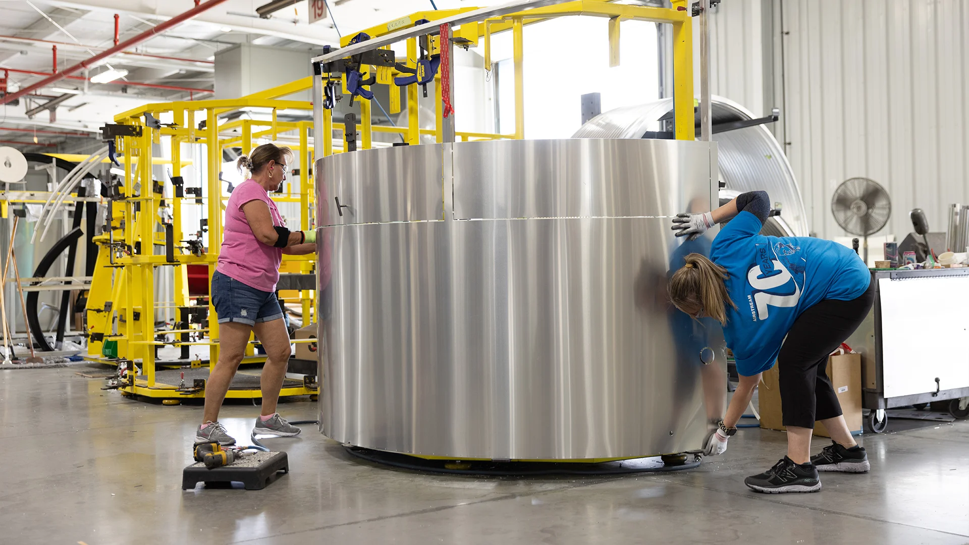 Two people work on a large roll of aluminum in an industrial setting, surrounded by machinery and tools.