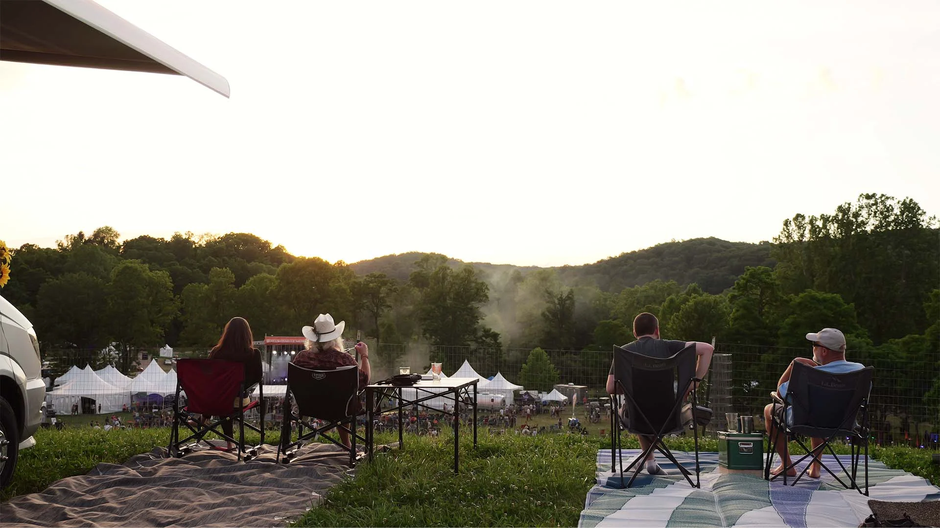 Four people sitting in camping chairs on a grassy hill overlooking a festival with white tents and a tree-covered background.