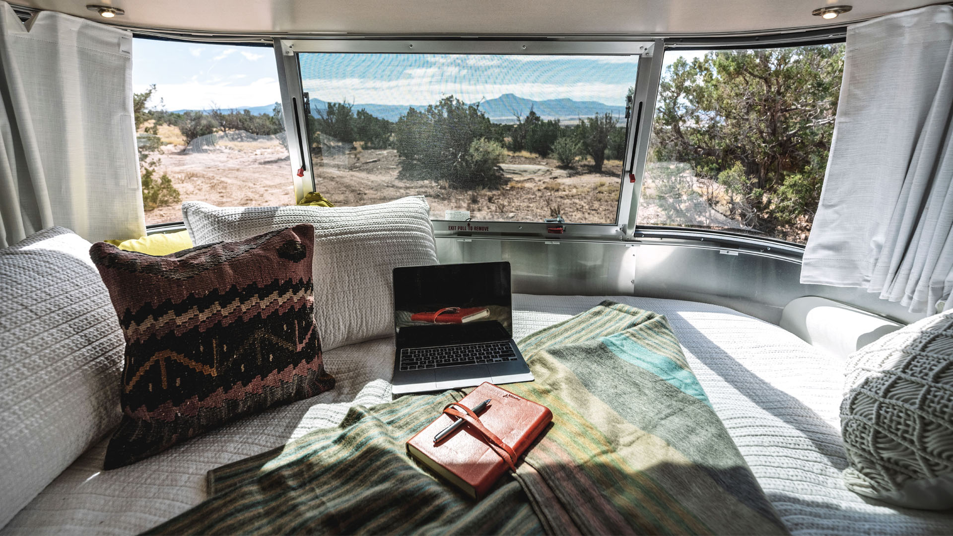 Cozy camper interior with pillows, a laptop, and a notebook, overlooking a sunny desert landscape through large windows.