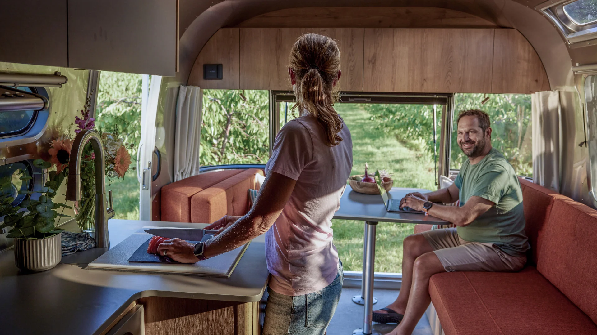 A person stands at a kitchen counter in a camper van, talking to a man seated at a dining table with a laptop. Sunlight fills the space.