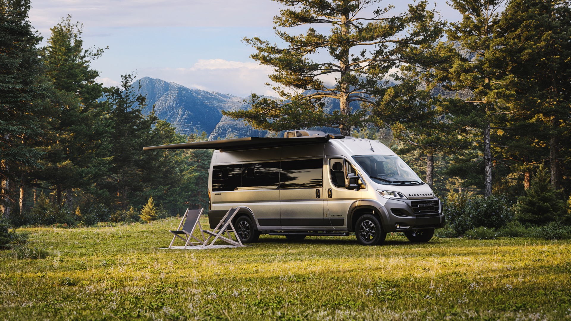 Camper van with an awning and chairs on a grassy field, surrounded by trees and mountains in the background.