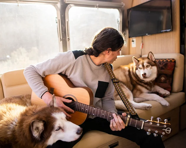 Man playing guitar inside an Airstream with two huskies, one sitting beside him and the other lounging on the couch, sunlight streaming through windows.