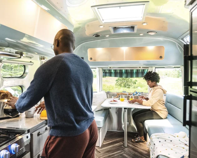 A man makes breakfast in the kitchen area of a camper van while a woman sits at a dinette table with food and drinks, looking at her phone. Sunlight streams in through the windows, creating a cozy atmosphere.