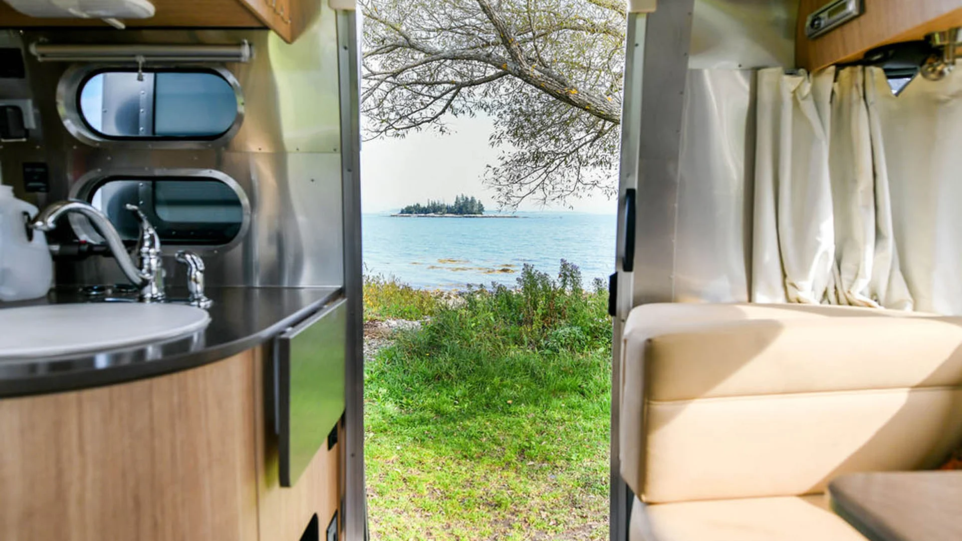 View from inside an RV shows a sink, tan seating, and a curtain, looking out to a grassy area by a lake or ocean with a small island and trees in the distance under a partly cloudy sky.