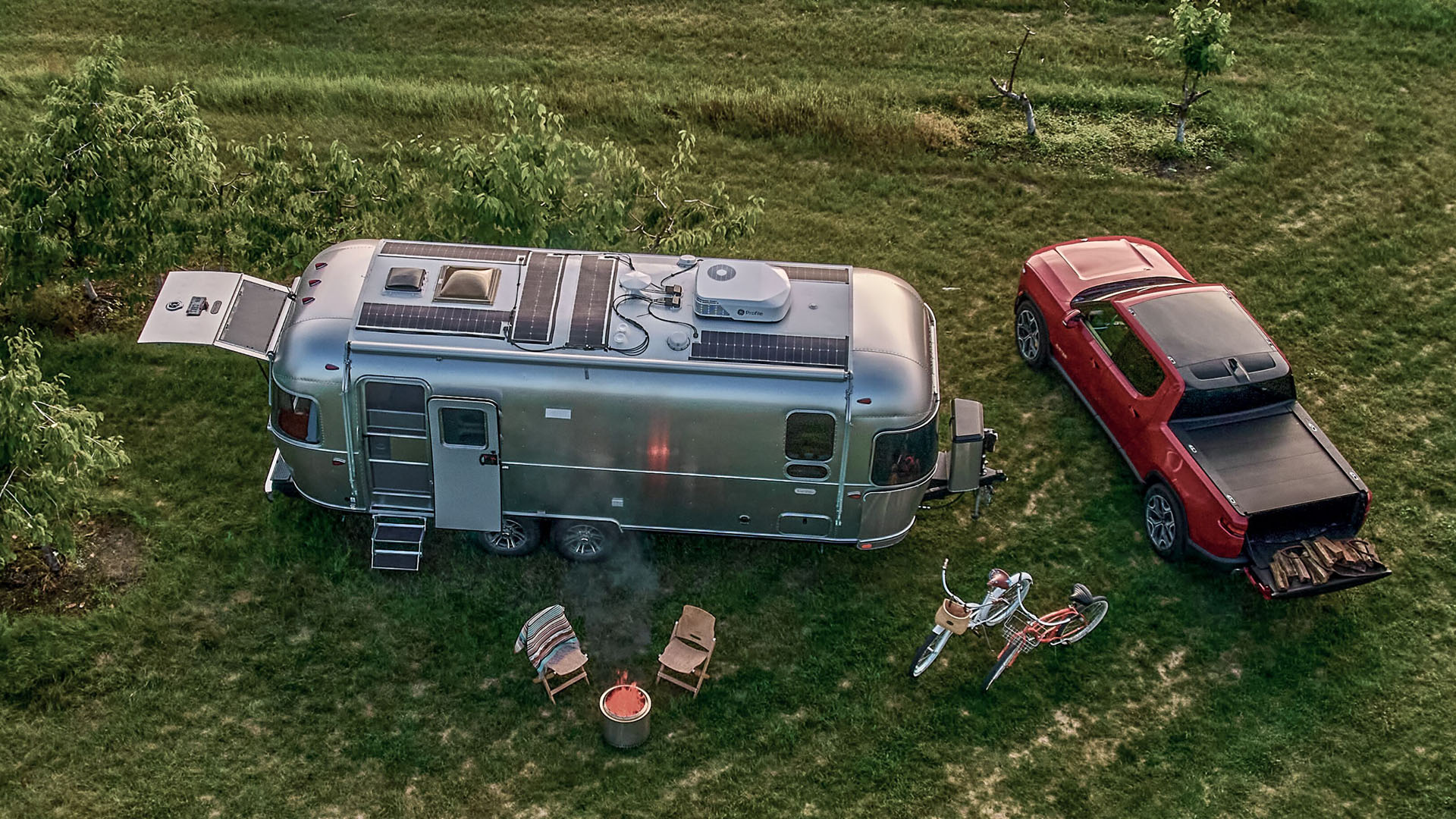 Aerial view of a silver RV and red pickup truck parked on grass, with two chairs, a bicycle, and a fire pit beside them.