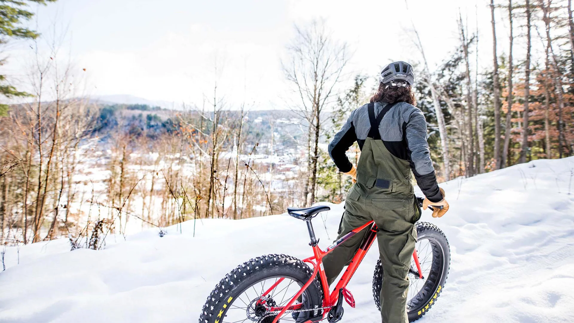 Person in winter gear standing with a red fat-tire bike on a snowy trail, overlooking a scenic valley with bare trees and distant hills.