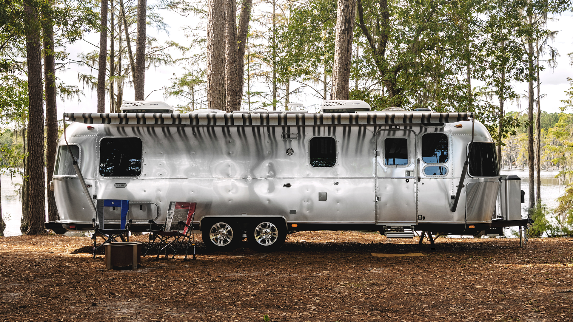 A silver Airstream trailer is parked among tall trees at a lakeside campsite, with chairs and a table set up outside.