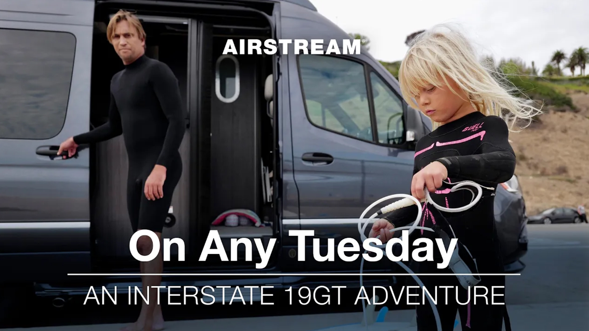 Child and adult in wetsuits near a van, preparing surf gear. Text reads, "Airstream On Any Tuesday: An Interstate 19GT Adventure."