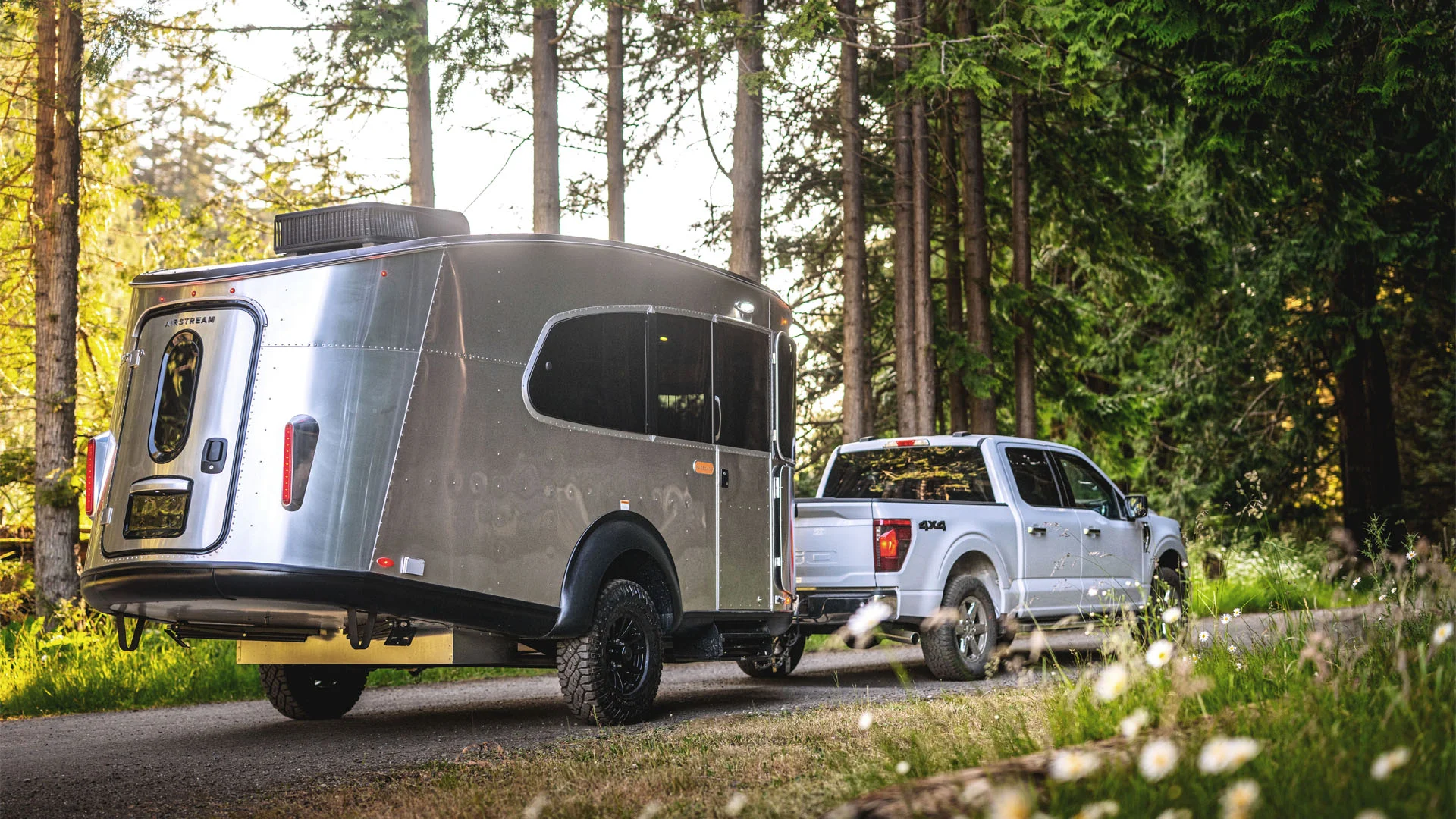 A silver pickup truck tows a shiny, modern travel trailer down a paved road through a forest, with tall trees and sunlight filtering through the branches in the background.
