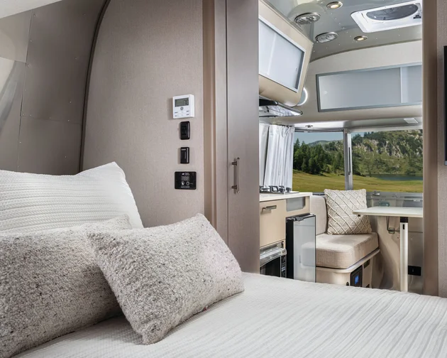 Interior of a modern RV with a neatly made bed in the foreground, a small kitchen area, a dining seat, and large windows showing a scenic outdoor view of grass and trees.