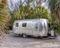 A shiny silver travel trailer parked among palm trees on a sandy area, with a clear blue sky peeking through the foliage.
