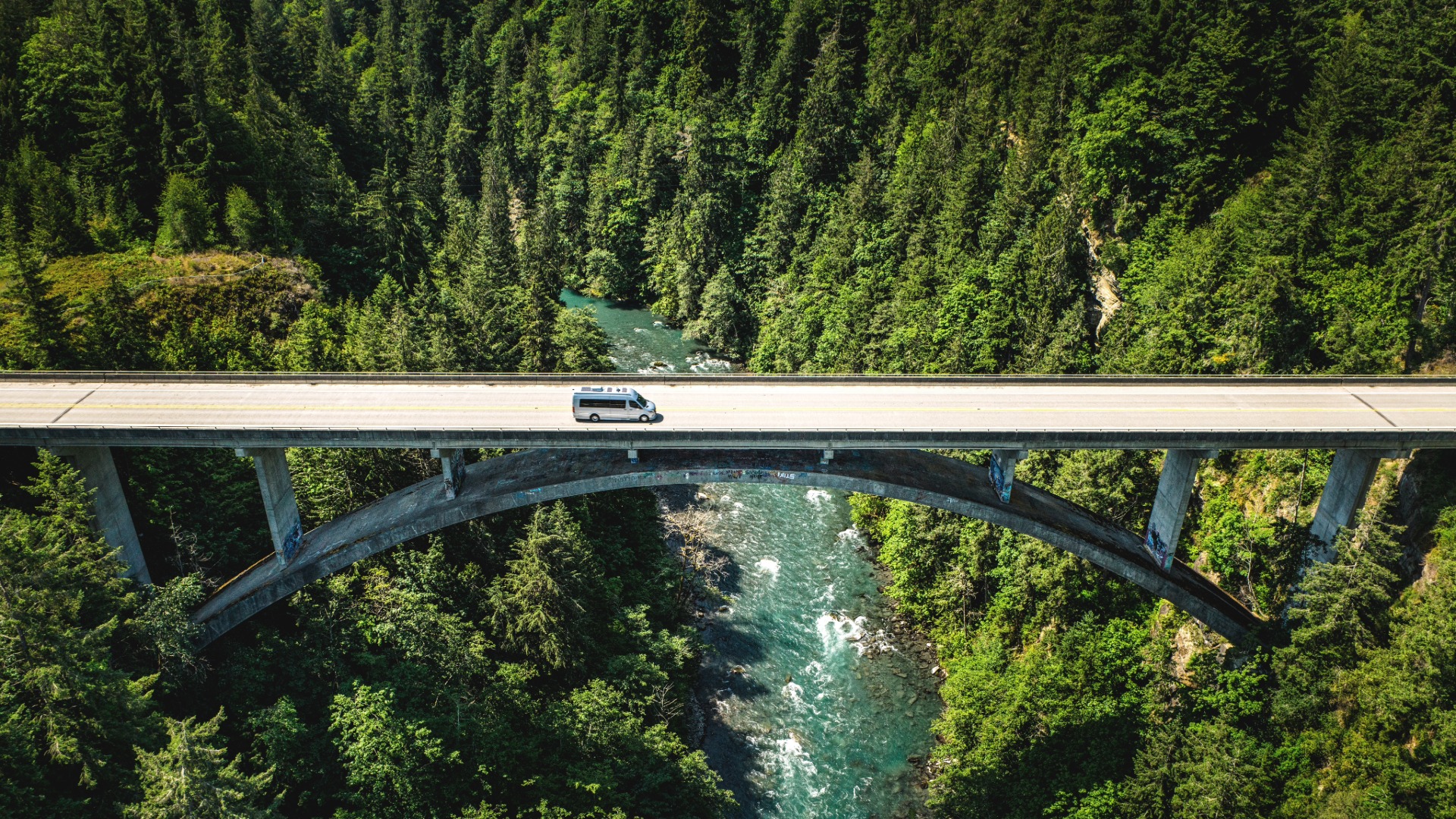 Aerial view of a van crossing a bridge over a lush forest and river, with dense green trees and clear water visible below.