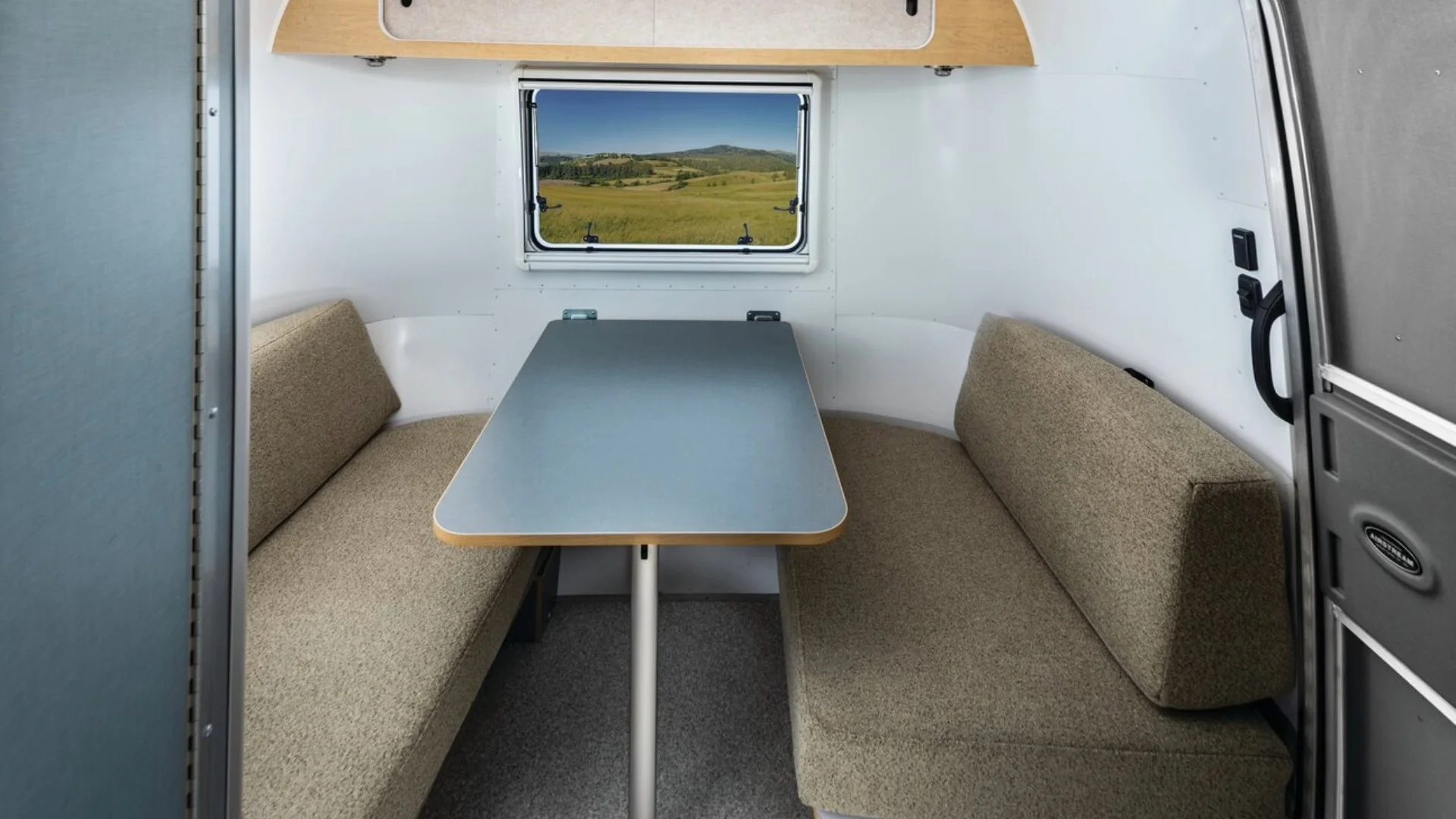 Interior of a camper features a compact dinette with a rectangular table and two cushioned benches. A window reveals a scenic landscape view.