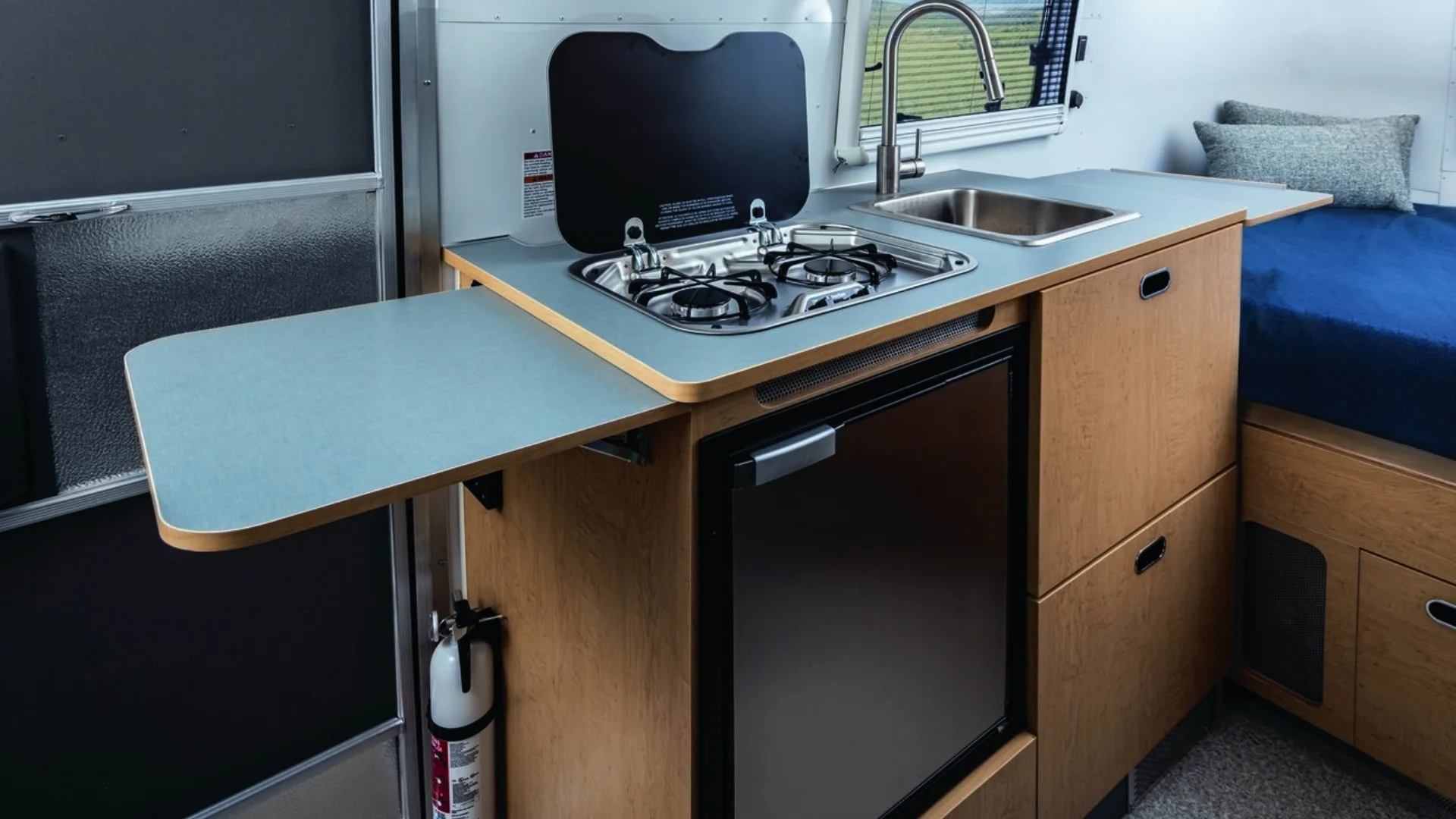 Compact RV kitchen area with a stove, sink, mini fridge, and fold-out counter, featuring wooden cabinetry and a small bed with cushions.
