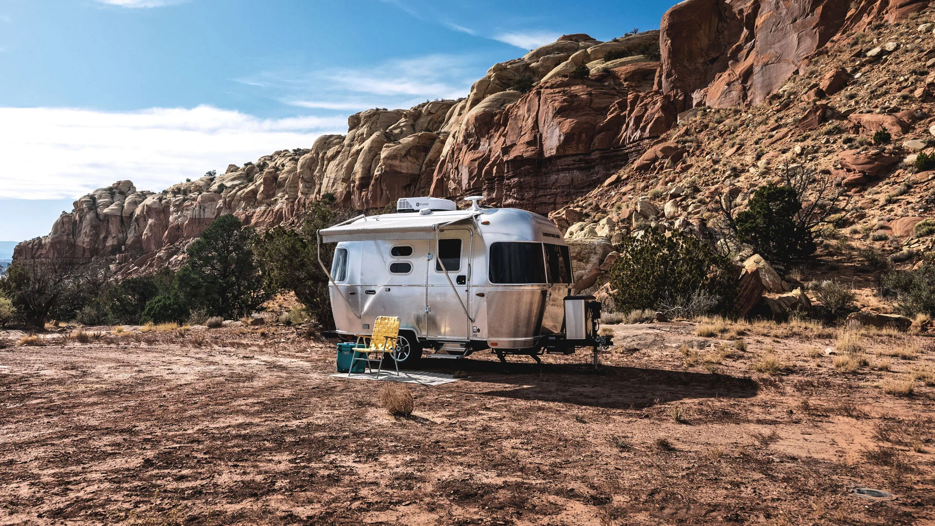 A silver Airstream trailer is parked on dry, rocky ground near large, rugged red rock cliffs. Camping chairs and a table are set up outside, under a blue sky with light clouds. Sparse desert vegetation surrounds the area.