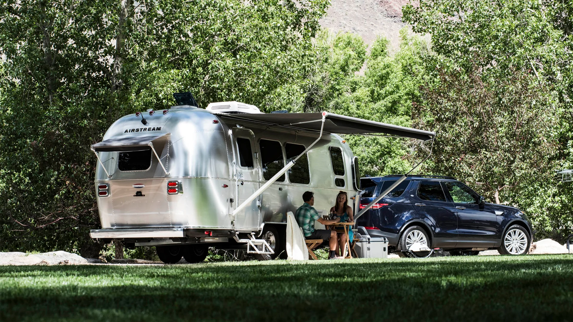 A silver Airstream trailer with its awning extended is parked next to a dark SUV in a green, wooded area. Two people sit at a table outside the trailer, enjoying a meal or conversation.