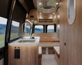 Interior of a modern camper van featuring wood cabinetry, a small sink, cushioned bench seats, overhead storage, and large windows with a scenic view of green hills outside.