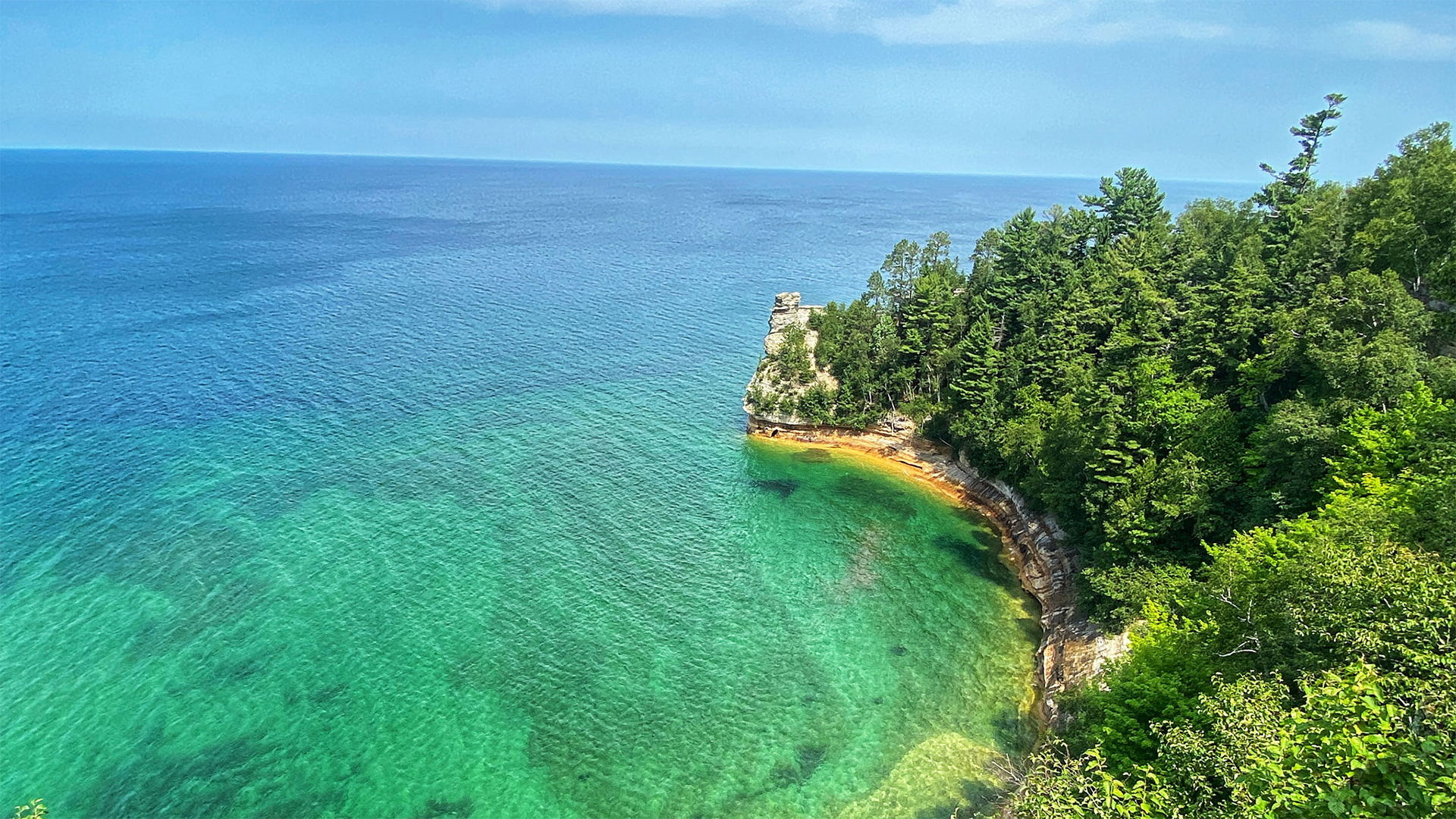 Aerial view of a clear turquoise bay surrounded by lush green cliffs and trees under a clear blue sky.