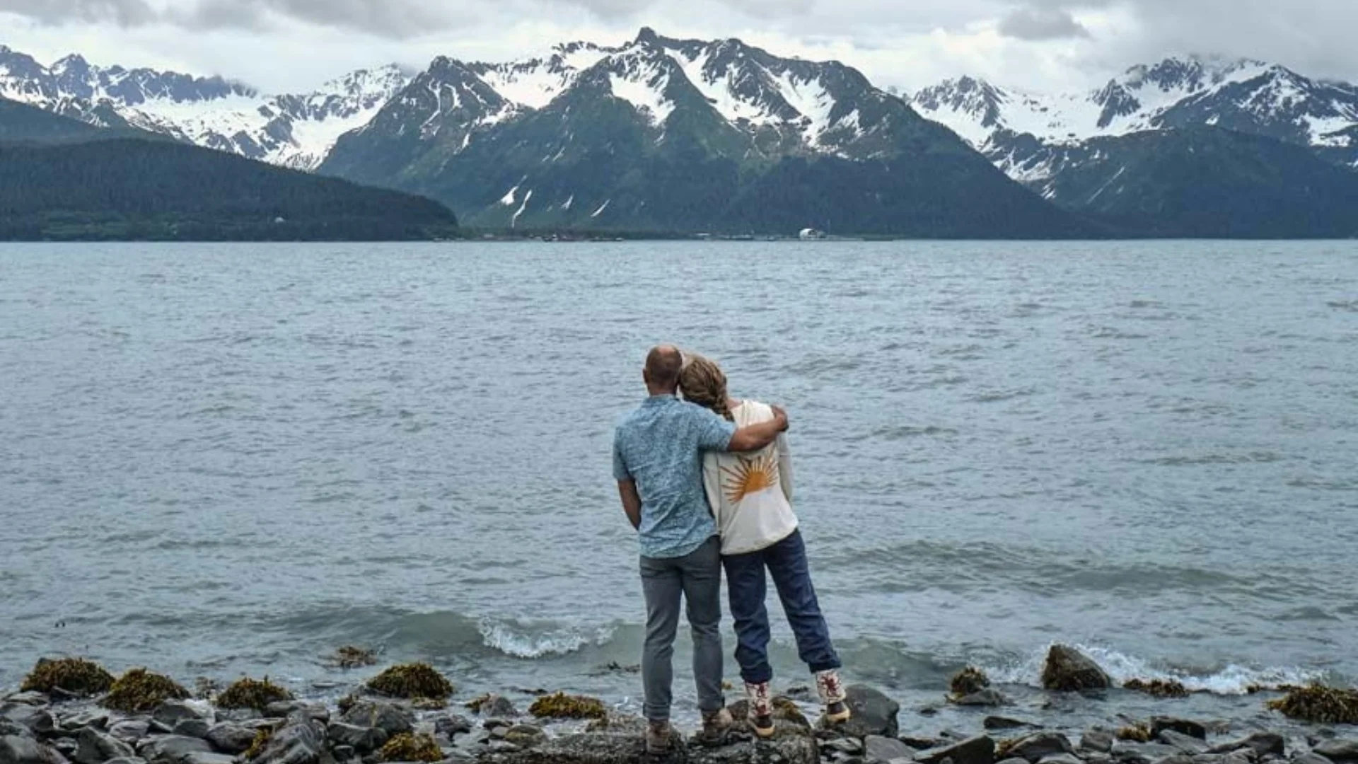 A couple stands embraced on a rocky shore, gazing at a mountain range across the water under a cloudy sky.