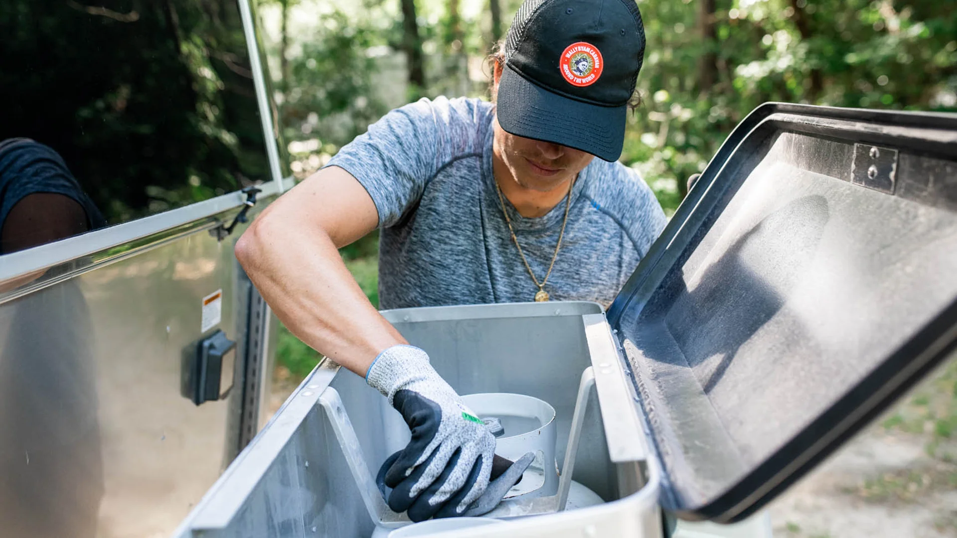 A person wearing a black cap, gray shirt, and gloves inspects or retrieves equipment from a gray container outdoors, with trees and sunlight in the background.