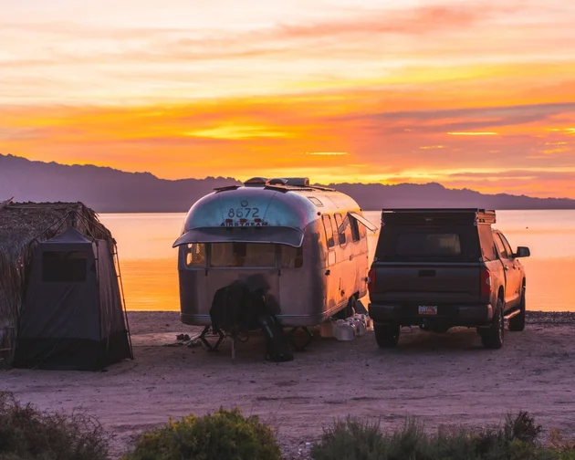 Airstream trailer and truck parked by a beach hut at sunset, overlooking a tranquil lake with mountains in the background.