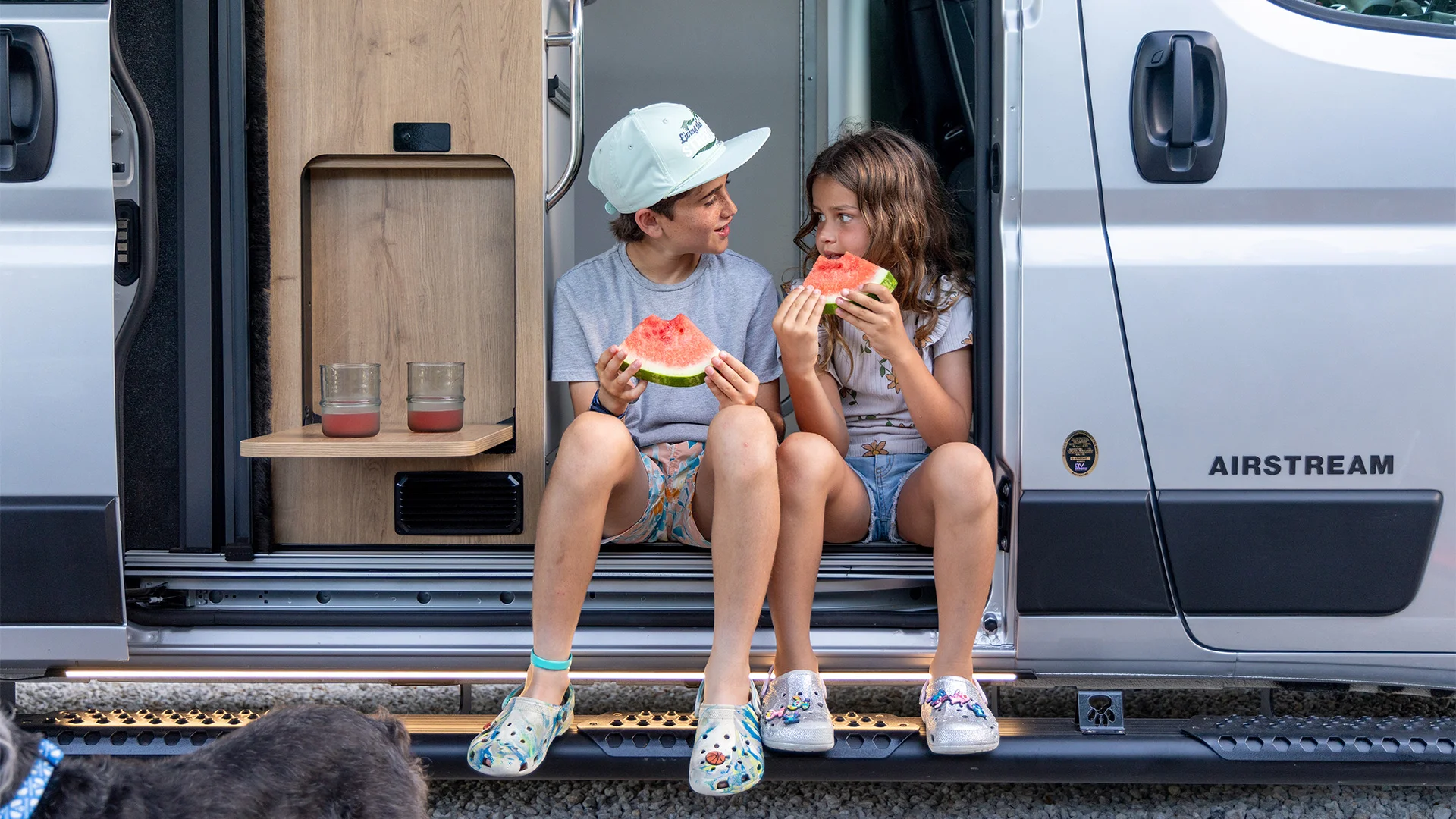 Two kids sit in a van doorway eating watermelon slices, with two glasses on a small shelf beside them. A dog's head is partially visible.