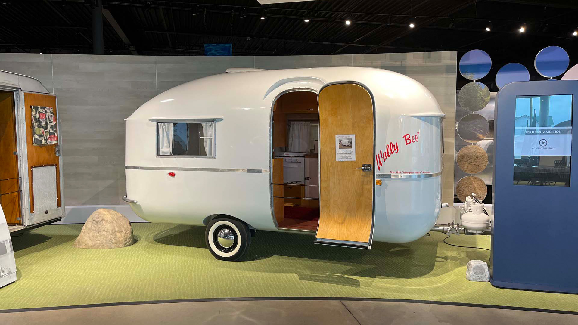 Vintage white travel trailer with wooden door open, revealing a small interior. It has "Molly Bee" text on the side and sits on a green carpet.