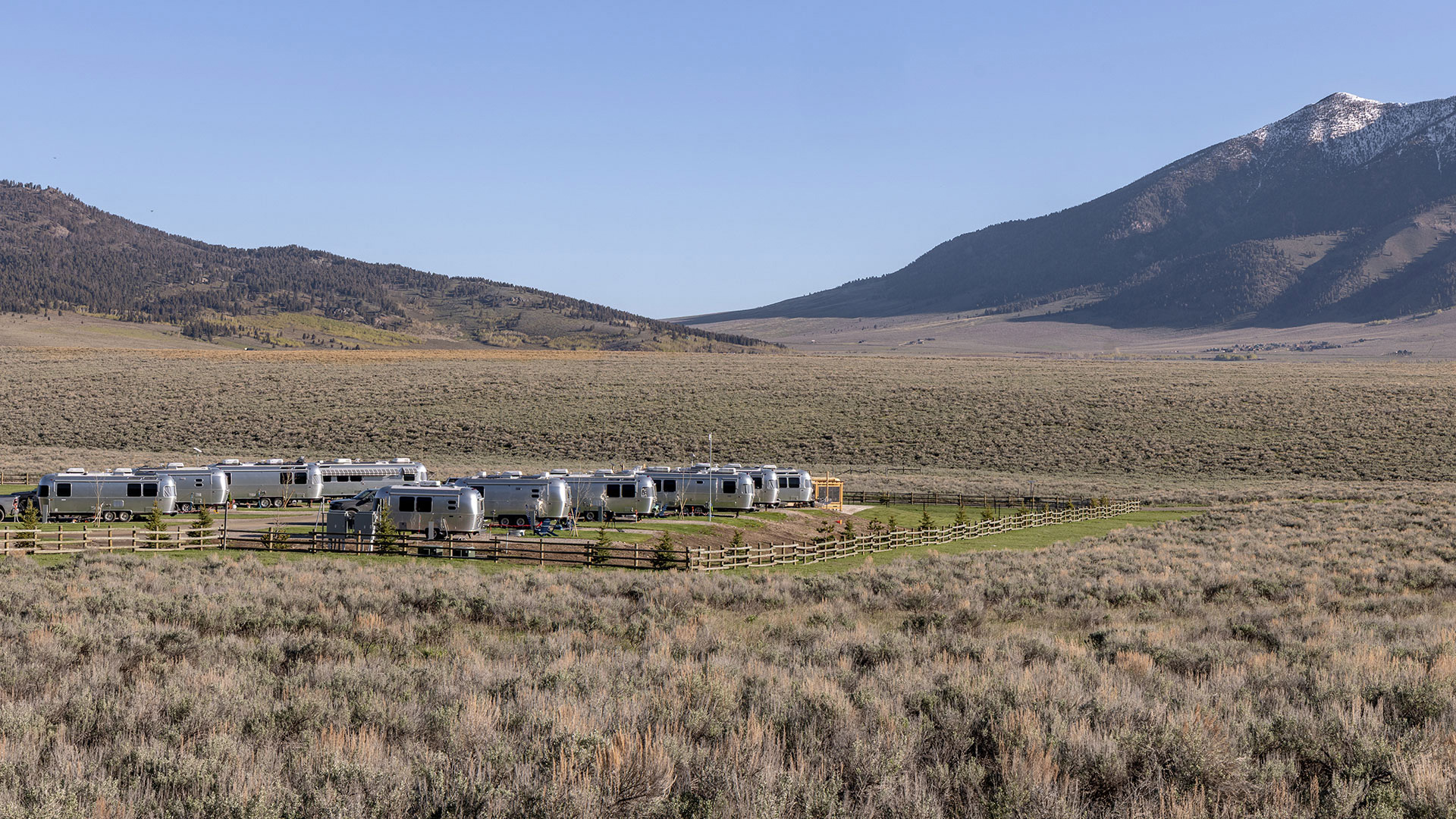 A row of RVs parked in a fenced area on a vast, grassy plain with mountains in the background under a clear blue sky.